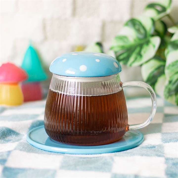 Clear mug with blue lid and base filled with tea on a light surface with colorful objects and plants in the background.