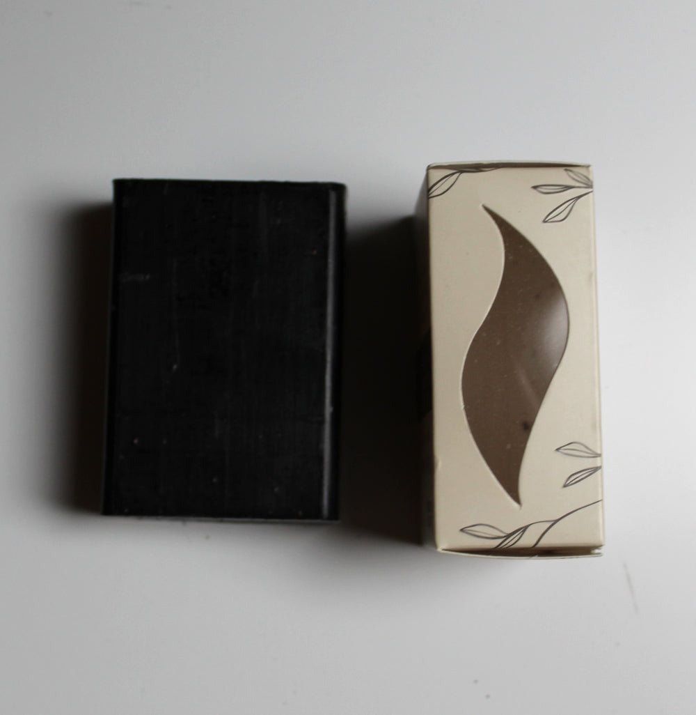 A rectangular black bar of soap stands next to its cream-colored packaging box with a brown leaf-shaped cutout window and delicate leaf line drawings.
