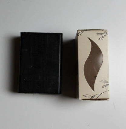 A rectangular black bar of soap stands next to its cream-colored packaging box with a brown leaf-shaped cutout window and delicate leaf line drawings.