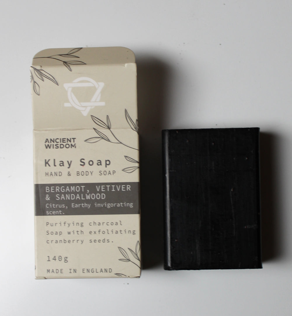 A 140g bar of Ancient Wisdom Klay Soap next to its beige packaging box, featuring a dark charcoal-colored soap bar and a box with botanical line drawings on a light gray background.