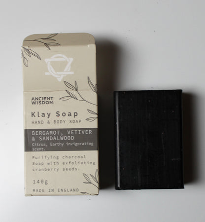 A 140g bar of Ancient Wisdom Klay Soap next to its beige packaging box, featuring a dark charcoal-colored soap bar and a box with botanical line drawings on a light gray background.