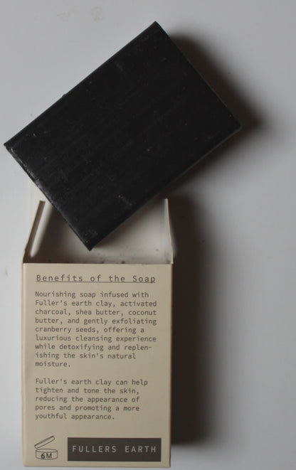 A black rectangular bar of soap resting on top of its open cream-colored cardboard box, which features printed text detailing the benefits of Fuller's Earth soap.
