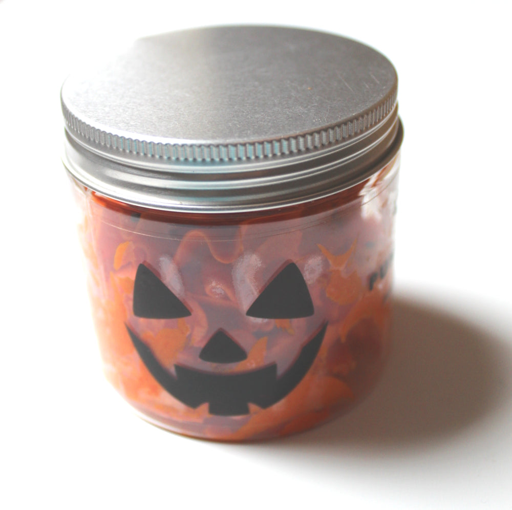 A small clear jar filled with pumpkin spiced whipped soap, featuring a black jack-o'-lantern face cutout on the front and sealed with a silver metal lid, set against a plain white background.