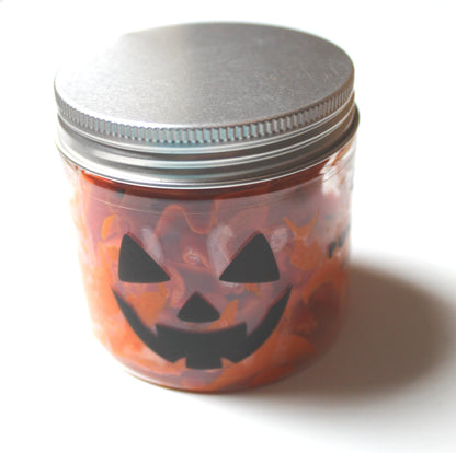 A small clear jar filled with pumpkin spiced whipped soap, featuring a black jack-o'-lantern face cutout on the front and sealed with a silver metal lid, set against a plain white background.