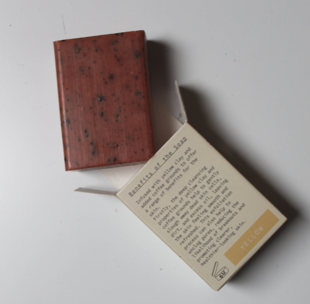 A rectangular bar of reddish-brown soap with dark flecks, shown next to its cream-colored paper packaging box on a plain white surface.
