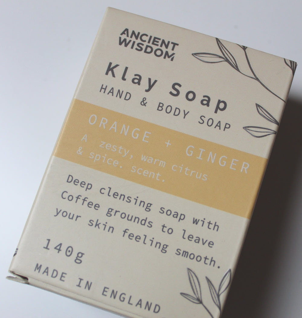 A 140g bar of Ancient Wisdom Klay Hand & Body Soap in the Orange + Ginger scent, shown in its beige cardboard packaging with a yellow-orange stripe.
