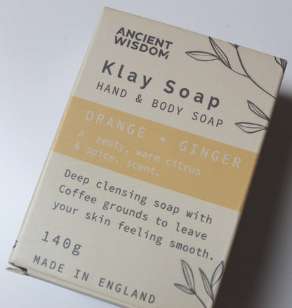 A 140g bar of Ancient Wisdom Klay Hand & Body Soap in the Orange + Ginger scent, shown in its beige cardboard packaging with a yellow-orange stripe.