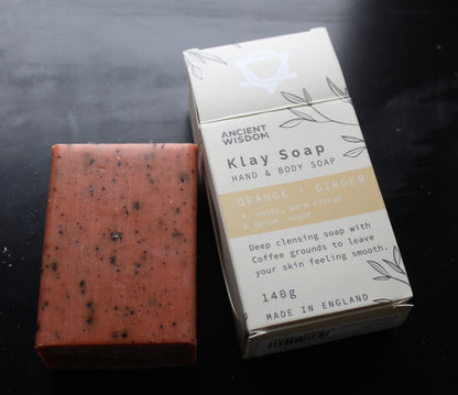 A rectangular bar of speckled orange soap next to its white and beige packaging box labeled 'Ancient Wisdom Klay Soap Orange + Ginger'.