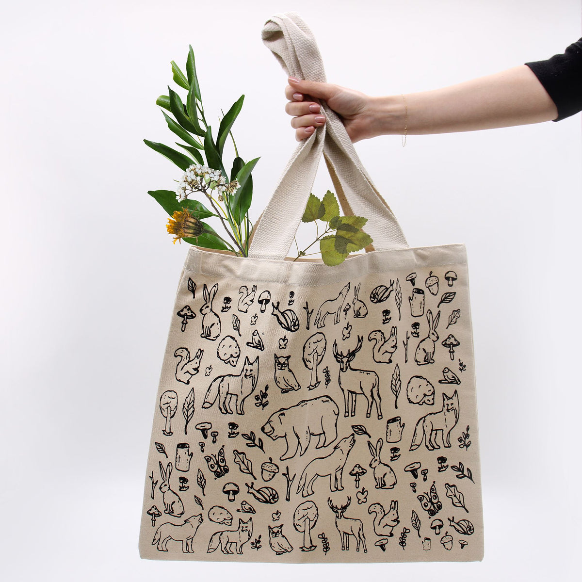 Beige tote bag with black animal illustrations held by a hand with greenery.