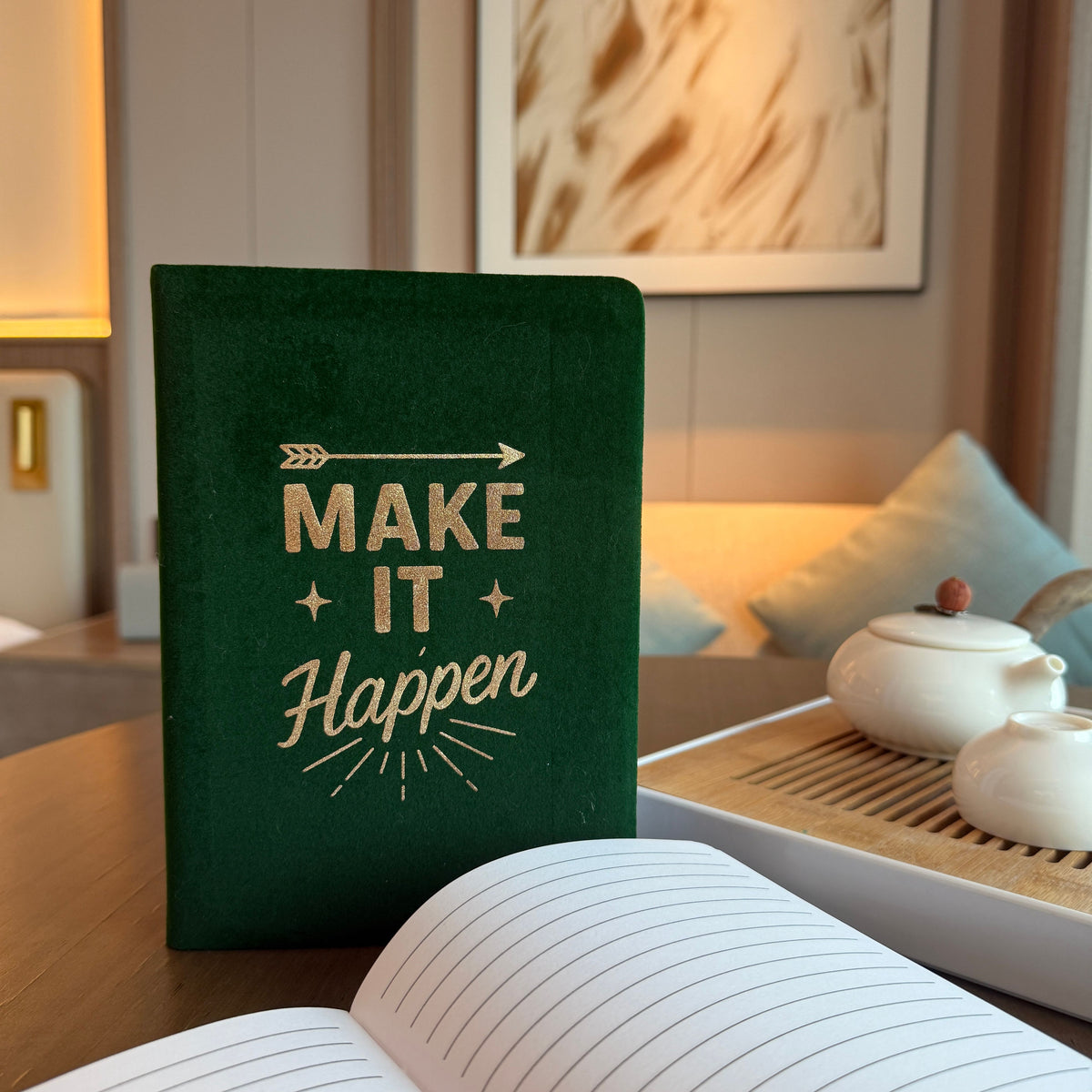 Green notebook with 'Make It Happen' text on a table with a tea set in the background