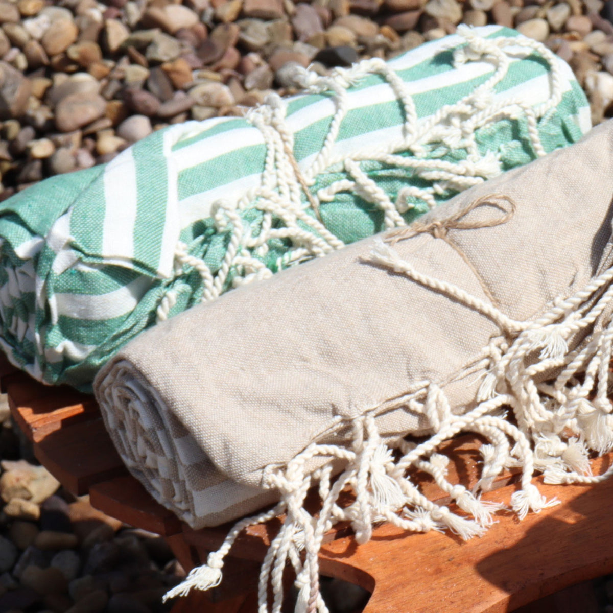 Two folded towels, one green and white patterned, the other beige with tassels, on a wooden surface with a stone background.