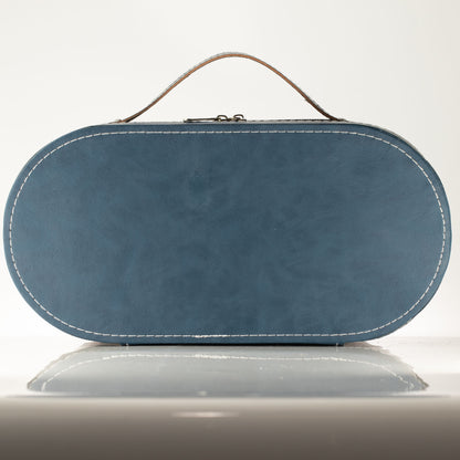 A blue oval leather clutch bag with a top handle, white contrast stitching, and a small metal clasp closure, shown on a reflective light gray surface against a white background.