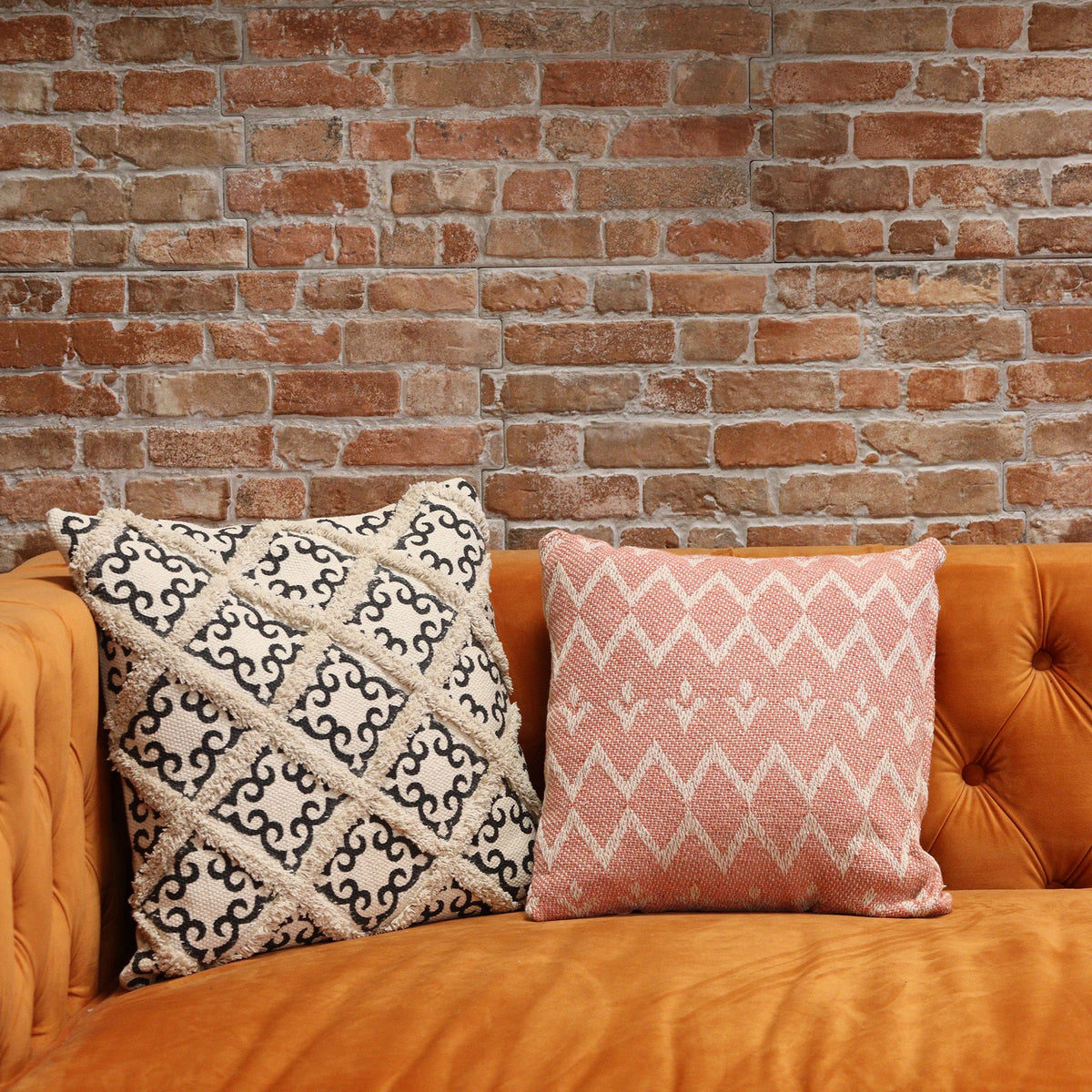 Two patterned pillows on a orange couch against a brick wall.