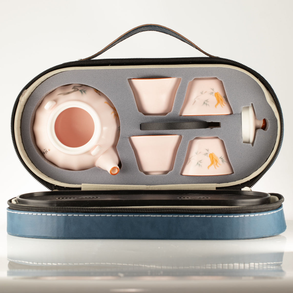 A ceramic tea set with a teapot, four cups, and a lid, arranged inside an open zippered hard case. The tea set has a pale pink glaze with delicate hand-painted floral and bird motifs.