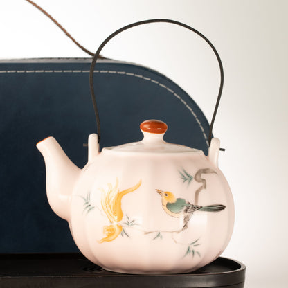 A ceramic teapot with a pale pink body, hand-painted birds and foliage, a black leather handle, and a small orange lid knob, shown against a neutral light background with a dark blue case behind it.
