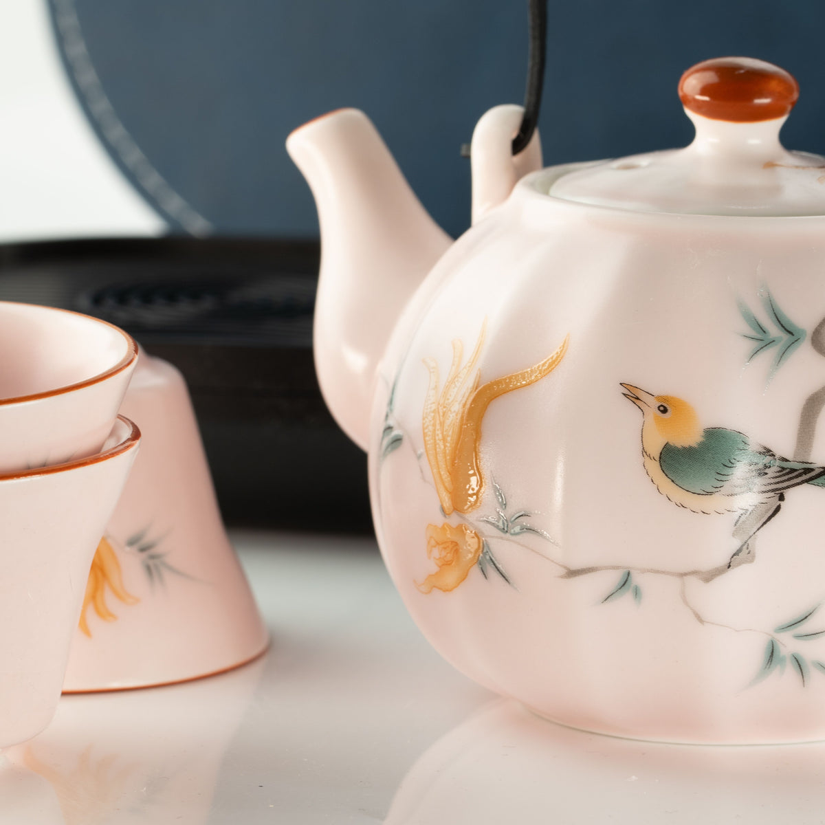 A close-up of a pale pink ceramic teapot with hand-painted bird and flower motifs, alongside matching teacups on a reflective white surface.