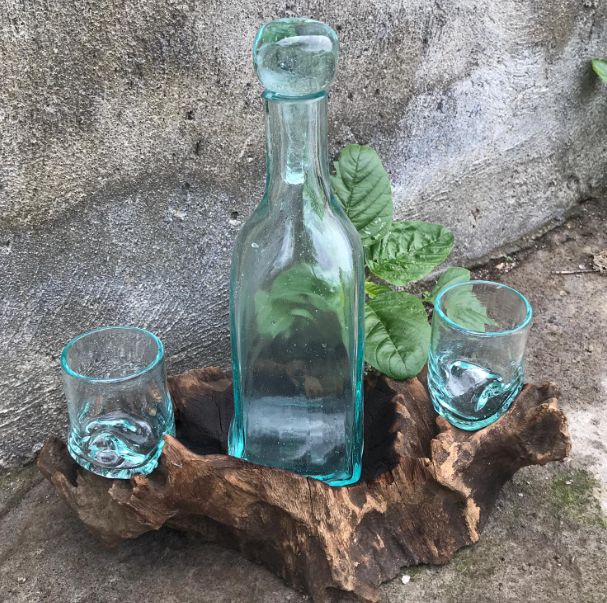 Molten glass whiskey set two glasses and a bottle on a piece of gamal wood root with greenery against a stone wall.